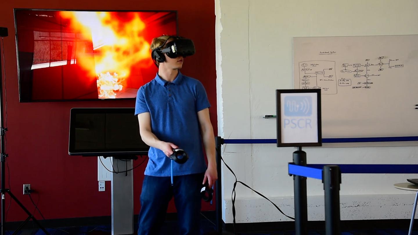 Jack Lewis demonstrates the use of a virtual reality headset and controllers with NIST's virtual office environment (shown on the screen behind him) in which first responders search for a body in a fire. Source: Burrus/NIST
